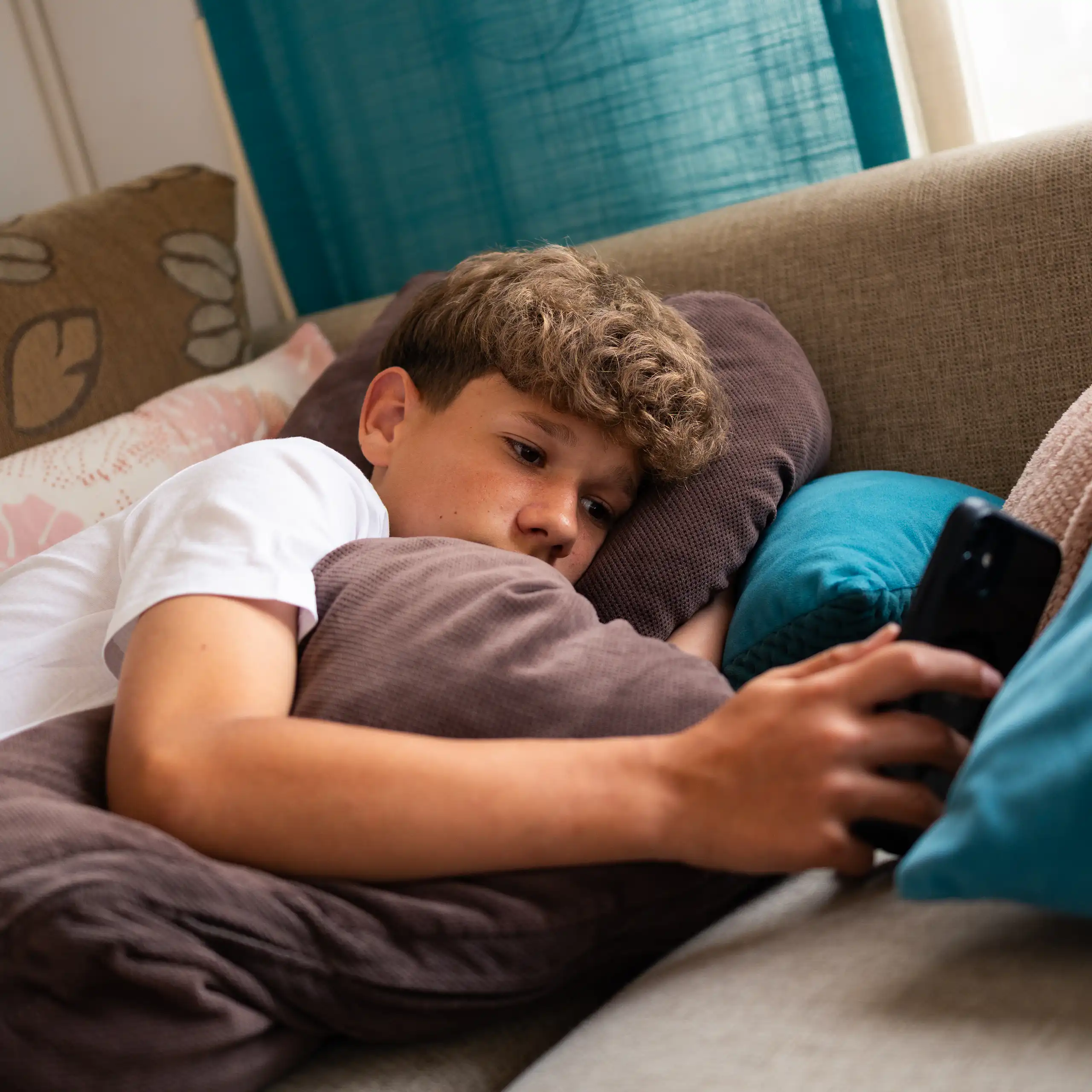 A teenage boy is relaxed on a couch, surrounded by soft cushions, focused on his smartphone.