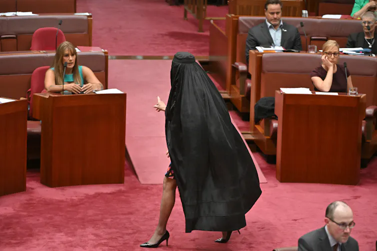 A woman in burqua, leg in stockings showing, in a parliamentary chamber.