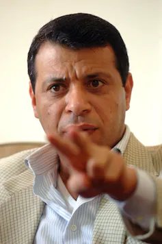 A man in a light-colored suit gestures to the camera.