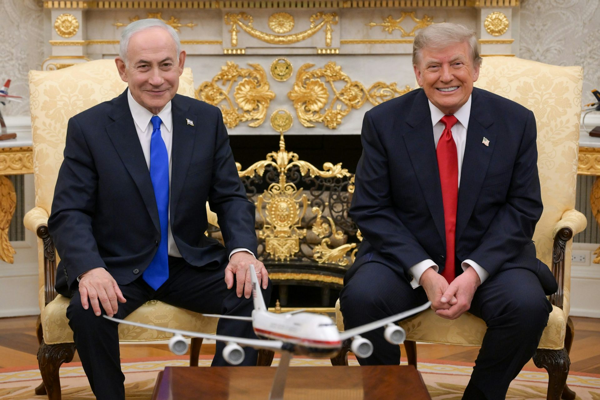 Two men in suits sit and smile in front of a table with a model airplane on it.