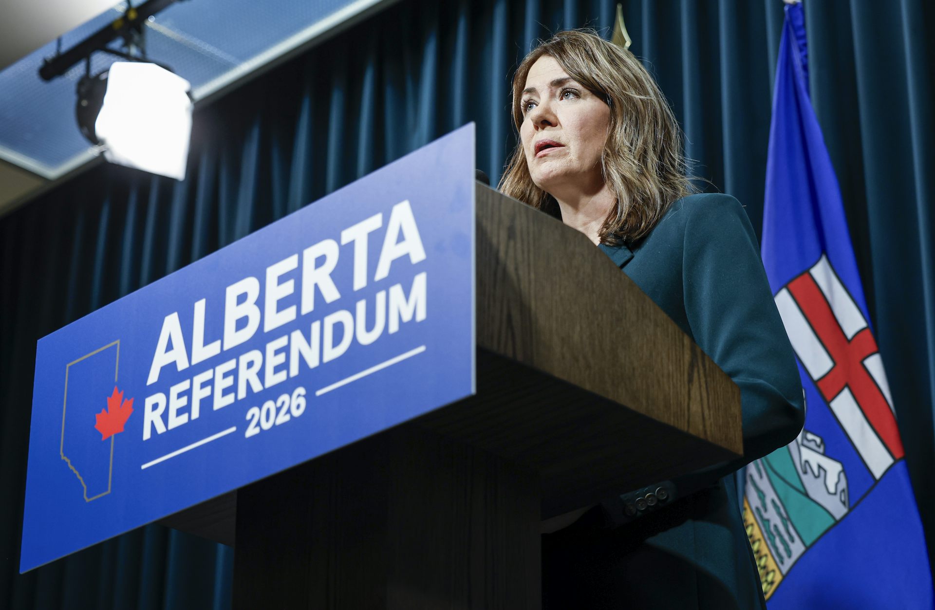 A dark-haired woman stands behind a podium with an 'Alberta Referendum' banner on it.