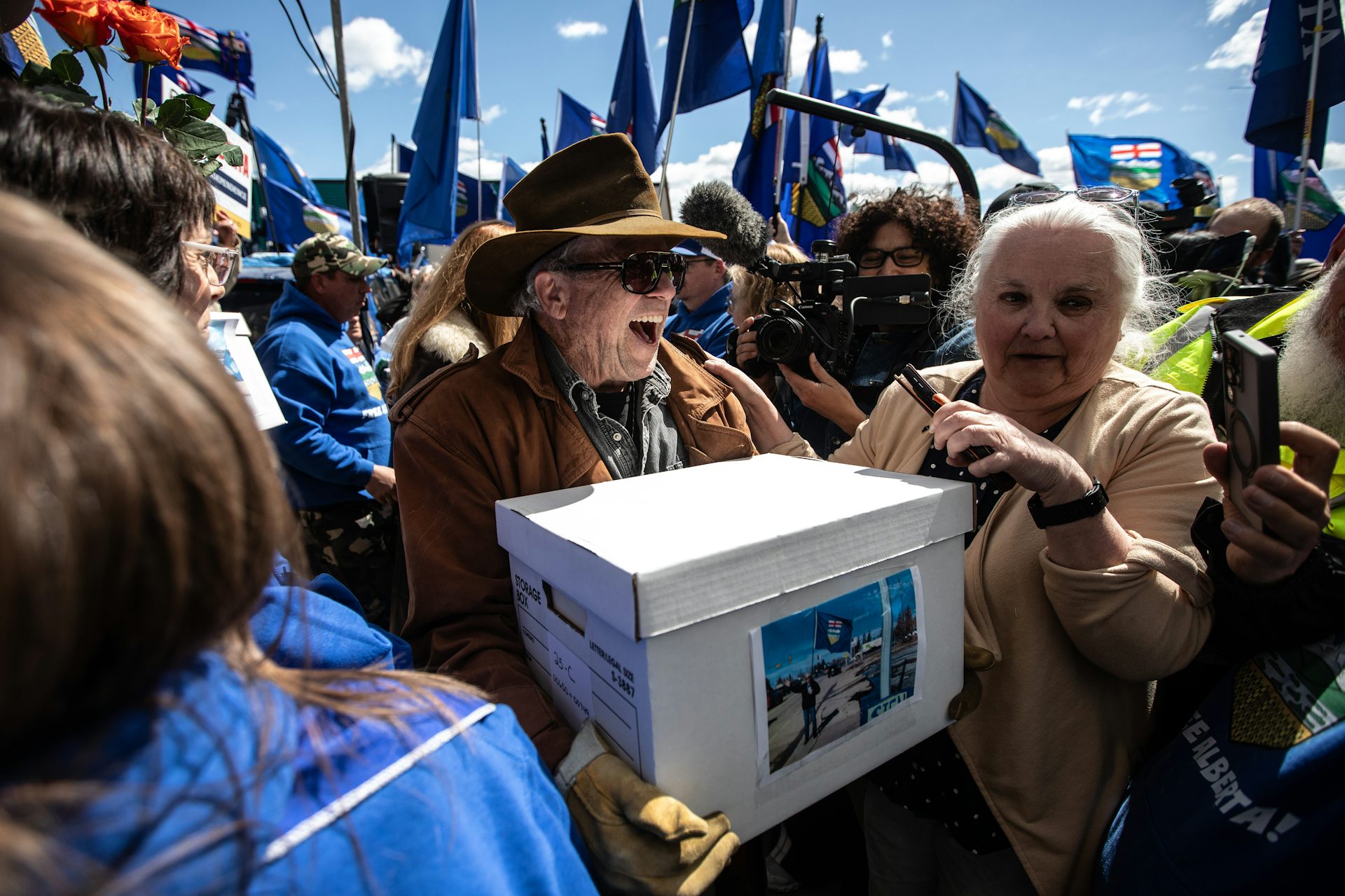 A shouting older man in a cowboy hat carries a cardboard box amid a crowd of other people.