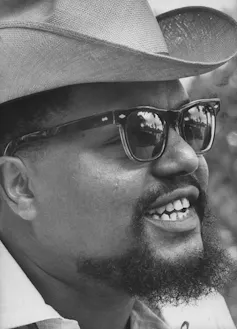 A Black man wearing 60s-style glasses, a shaggy beard and a cowboy hat is photographed from the side.