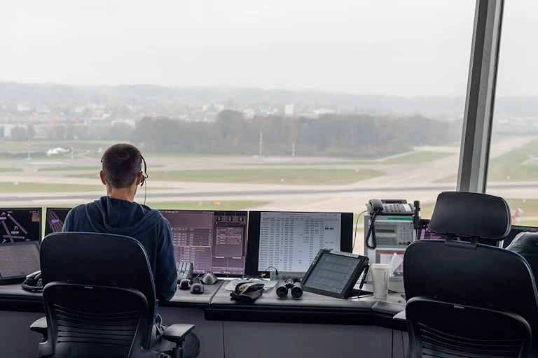 Person seen from behind sitting at a desk with computer screens, phone and binoculars, facing a big window looking onto an airport runway