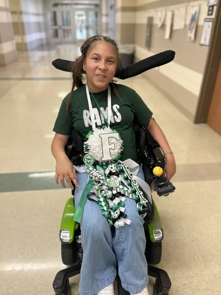 A girl with dark hair wears a green shirt that says Rams and has green, white and black ribbons on her lap. She sits in a wheelchair in an empty hallway.