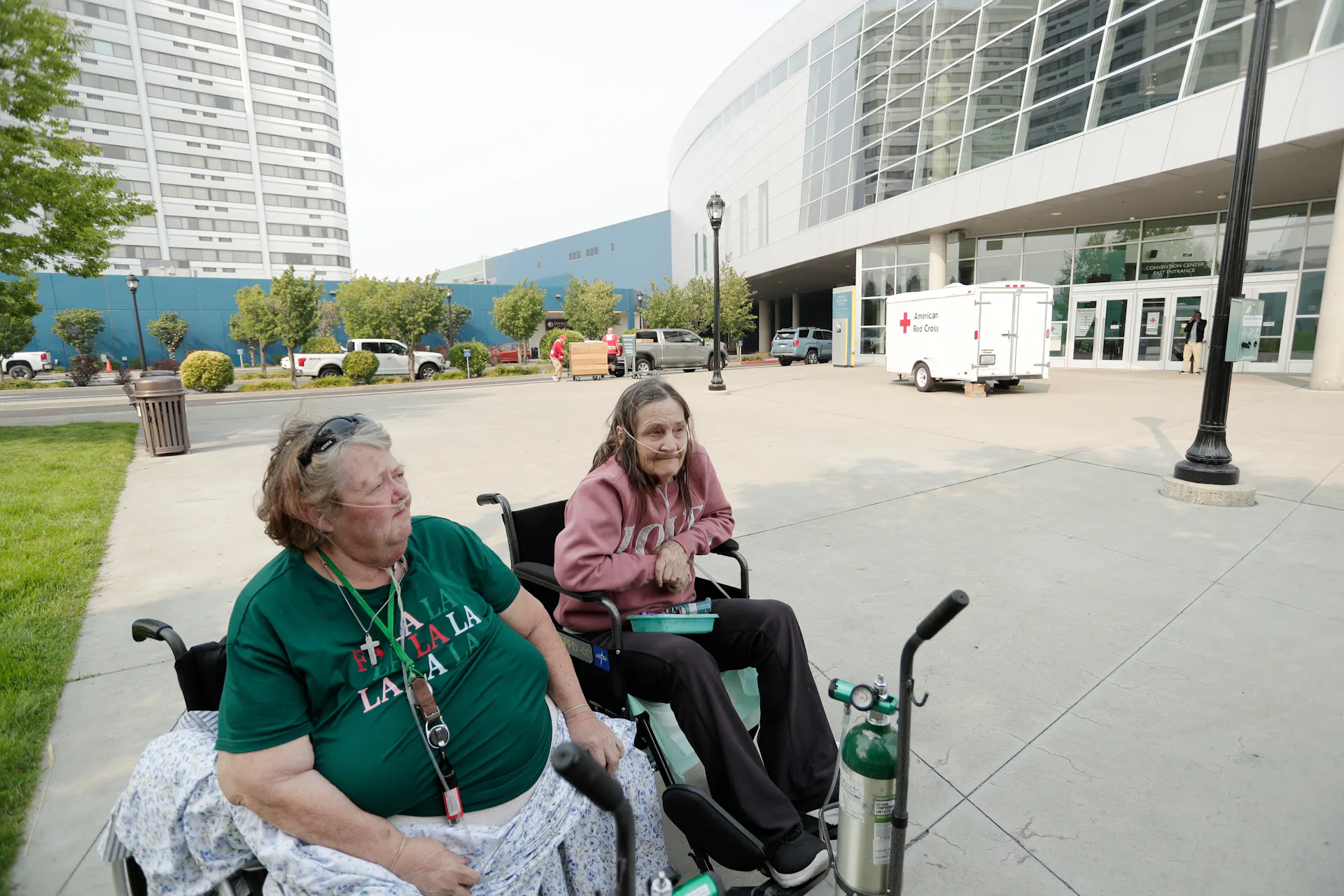 Two women sit in wheelchairs, one with an oxygen tank, outside a convention center. A Red Cross truck is in the background.