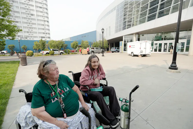 Two women sit in wheelchairs, one with an oxygen tank, outside a convention center. A Red Cross truck is in the background.