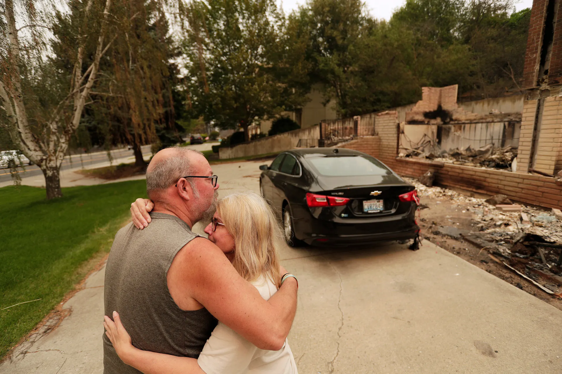 Two people hug next to a clean car parked next to the remains of a home.