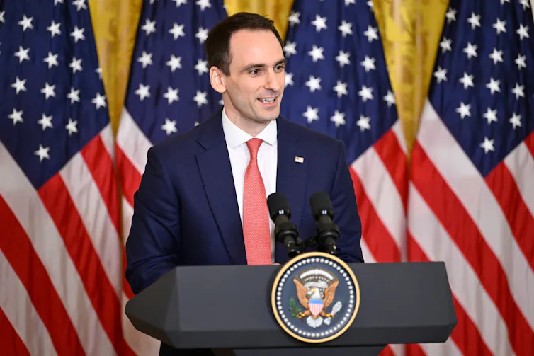 man in suit and tie stands at White House podium with American flags behind