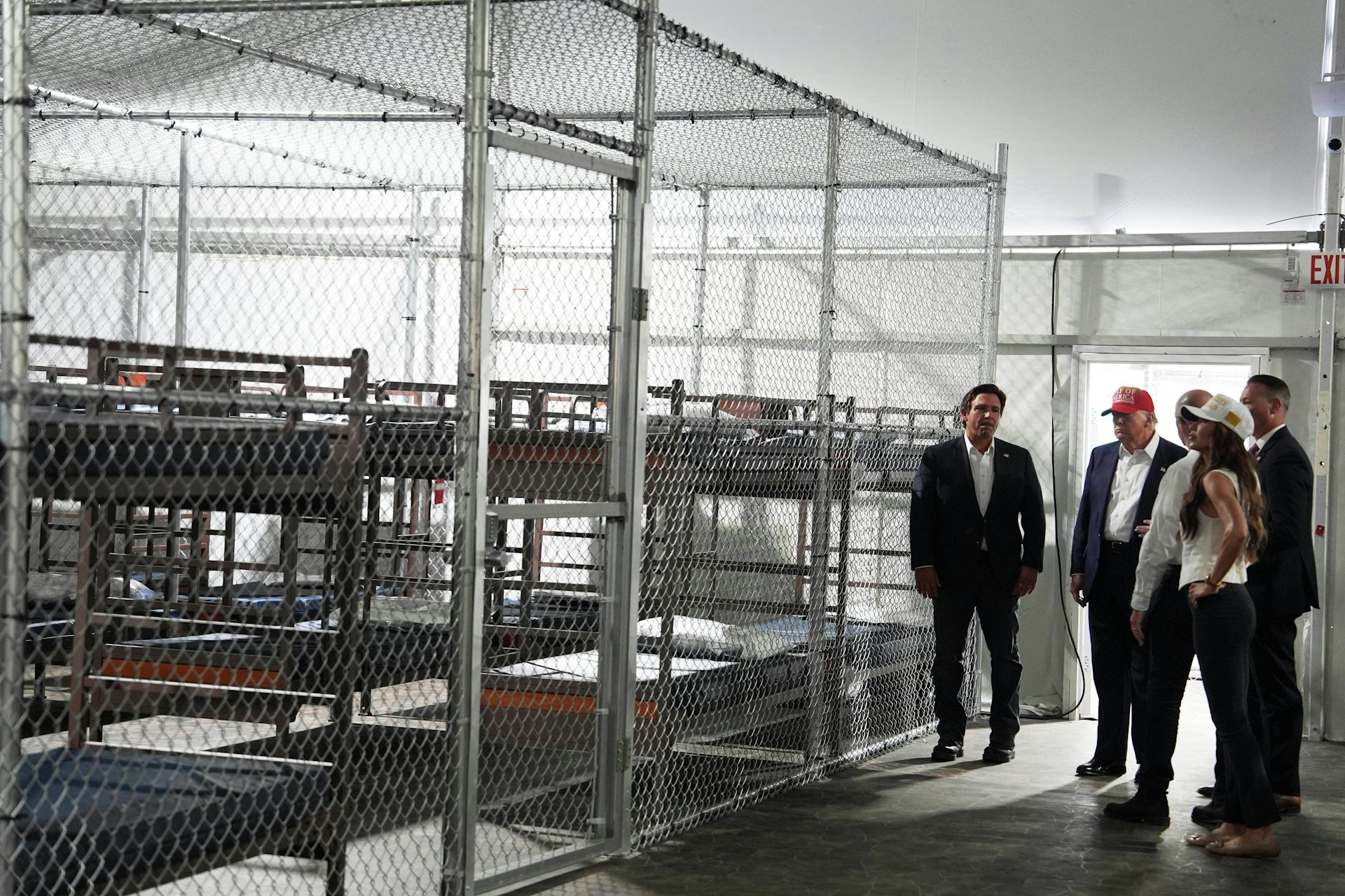 Donald Trump, Ron DeSantis and Kristi Noem overlook empty rows of bunk beds behind a tall chain-link fence inside a detention facility
