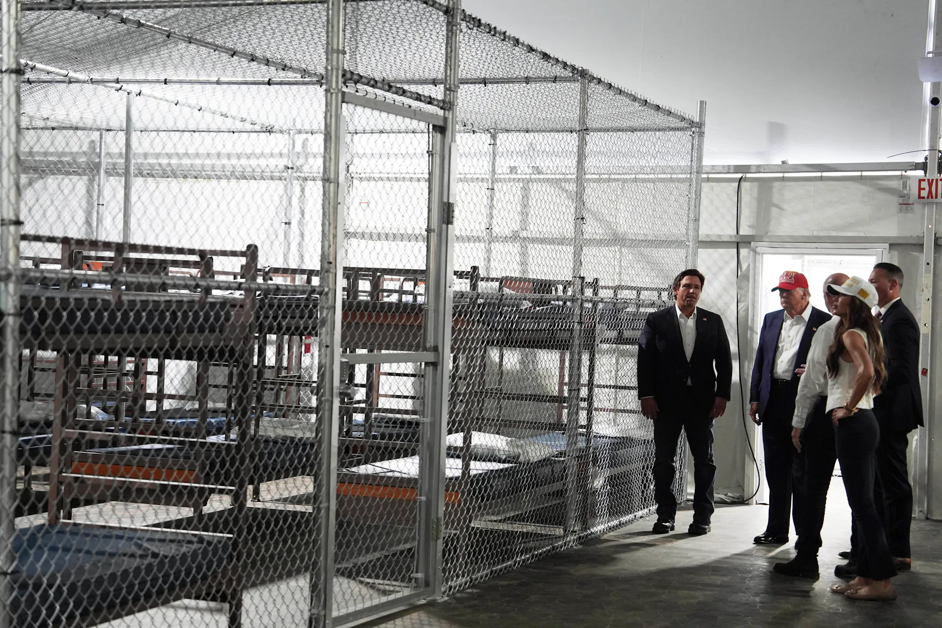 Donald Trump, Ron DeSantis and Kristi Noem overlook empty rows of bunk beds behind a tall chain-link fence inside a detention facility
