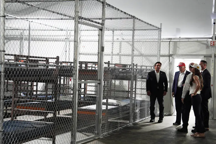 Donald Trump, Ron DeSantis and Kristi Noem overlook empty rows of bunk beds behind a tall chain-link fence inside a detention facility