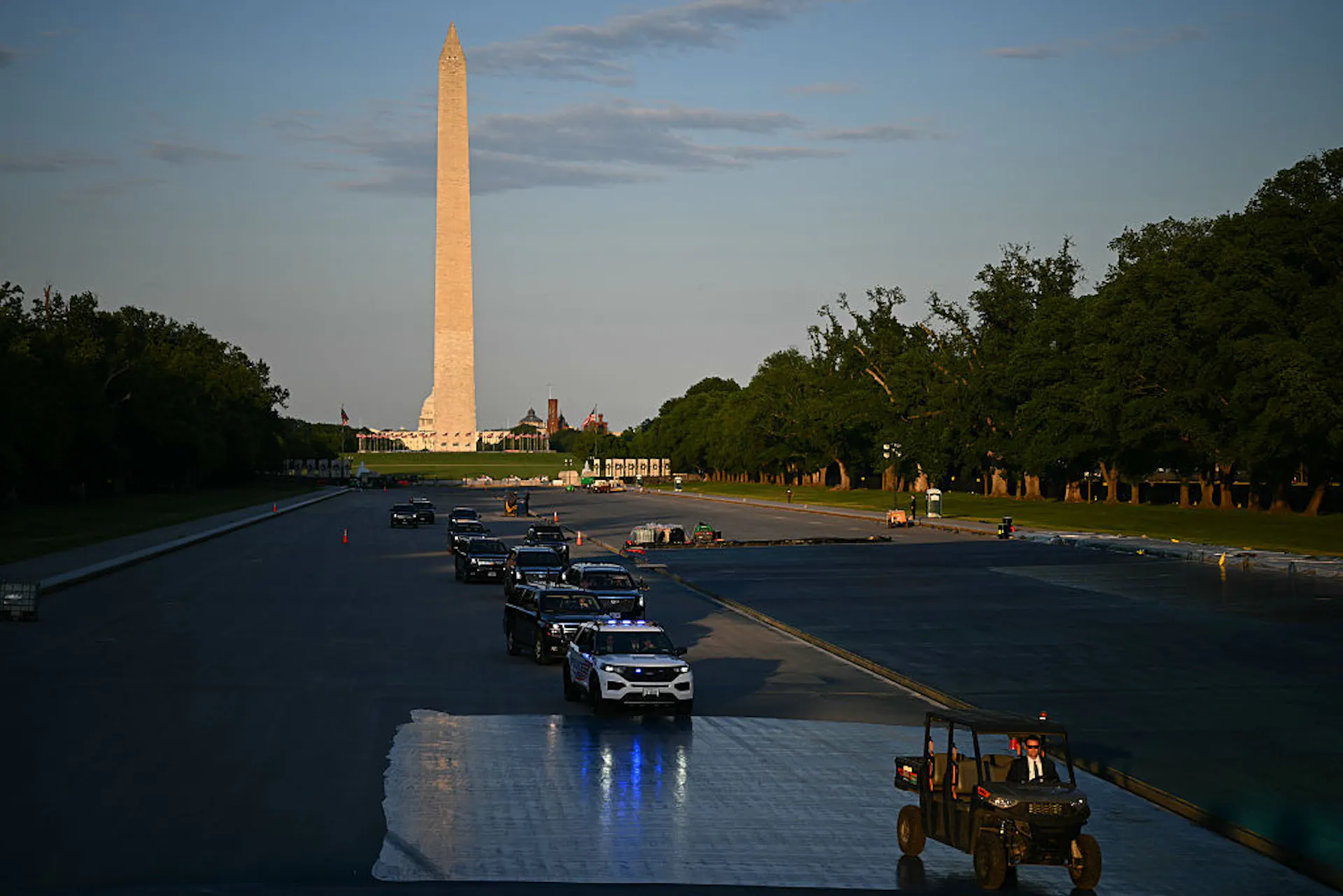 A convoy of vehicles drives over the floor of an emptied pool with a large white monument in the background.