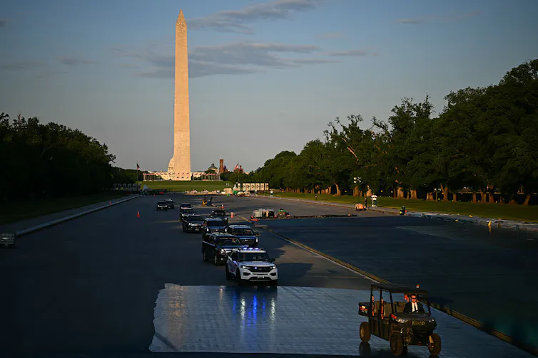 A convoy of vehicles drives over the floor of an emptied pool with a large white monument in the background.