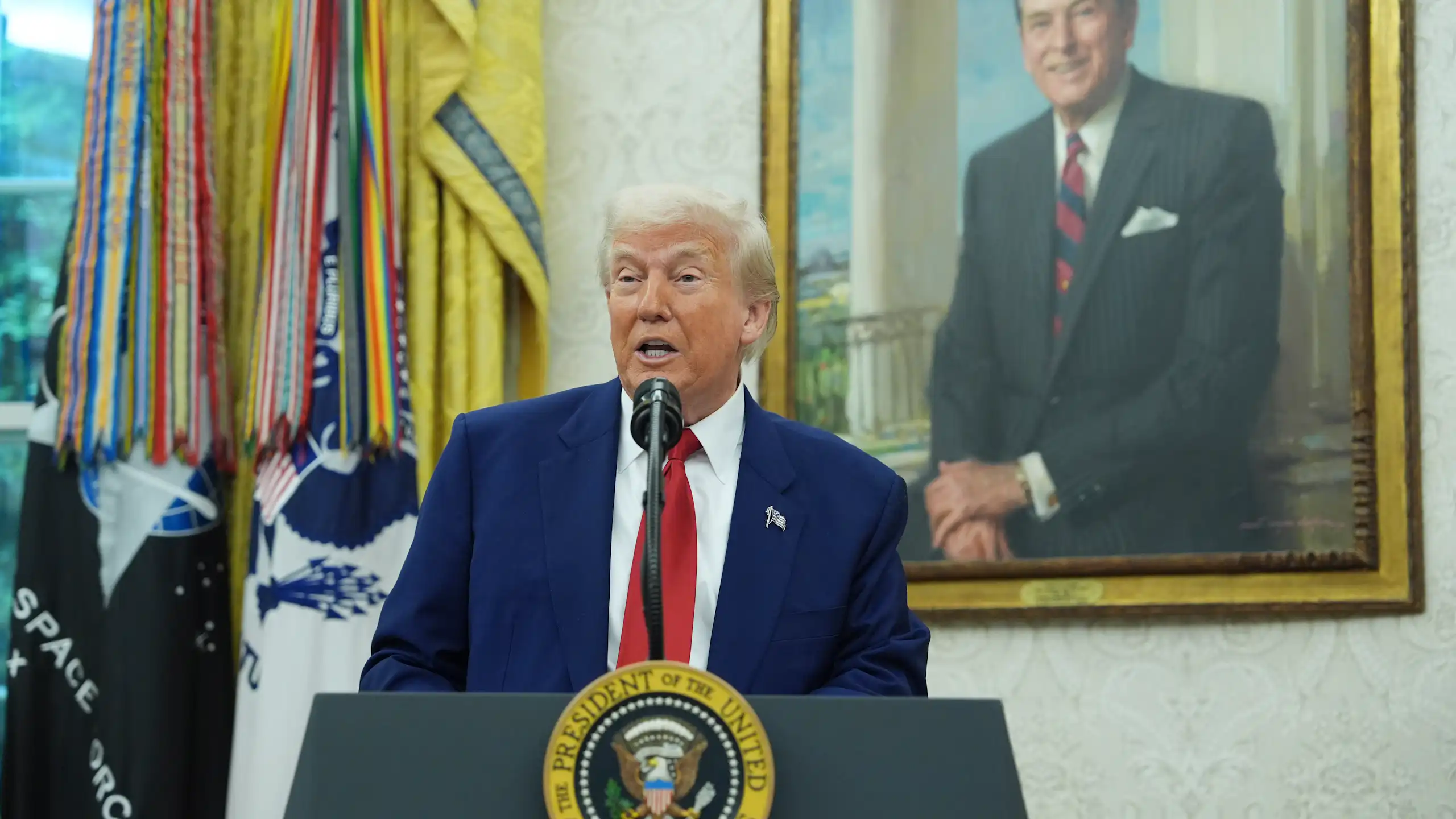 Donald Trump speaks at a lectern bearing the presidential seal with a painting of Ronald Reagan in the background