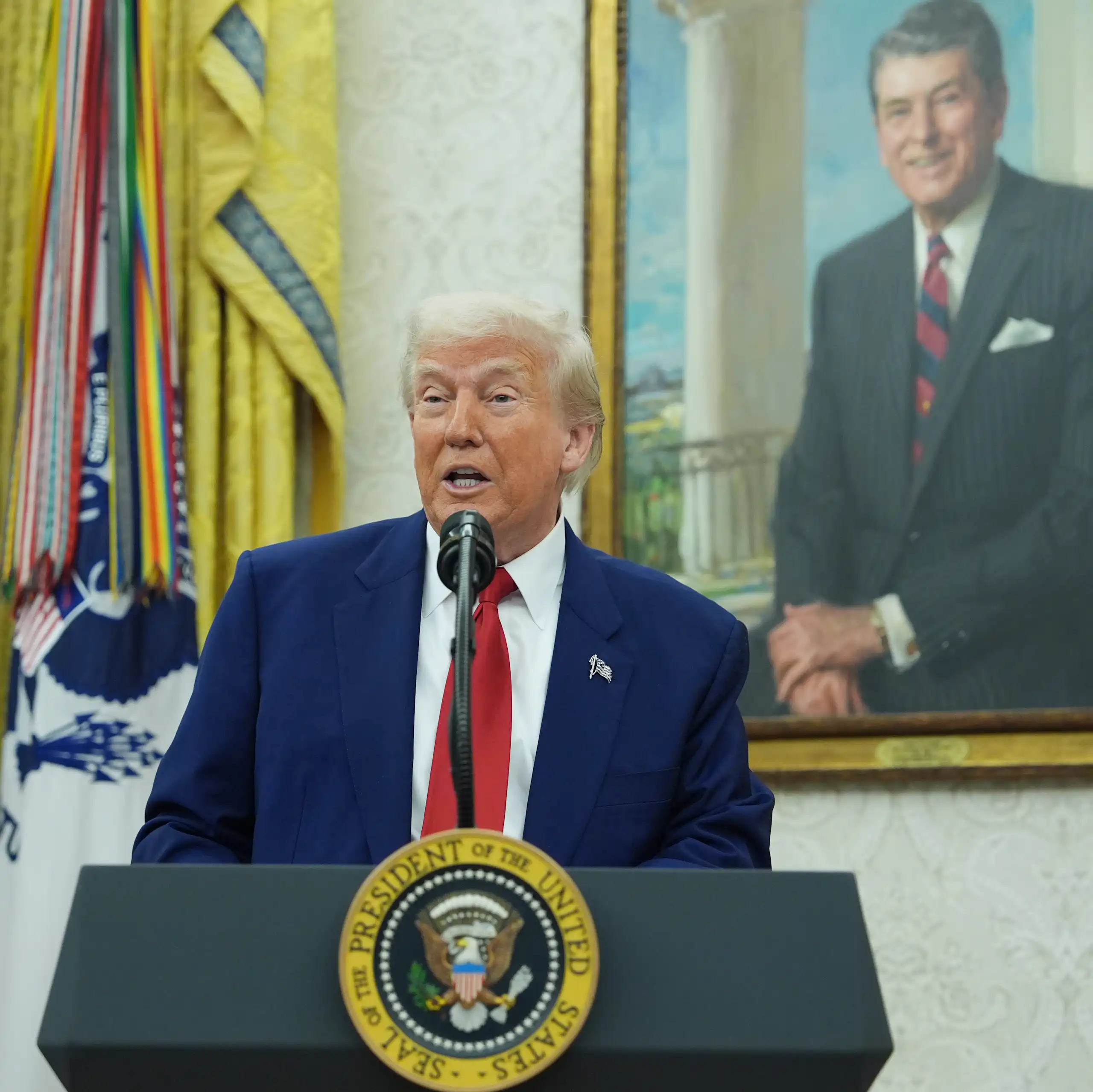 Donald Trump speaks at a lectern bearing the presidential seal with a painting of Ronald Reagan in the background