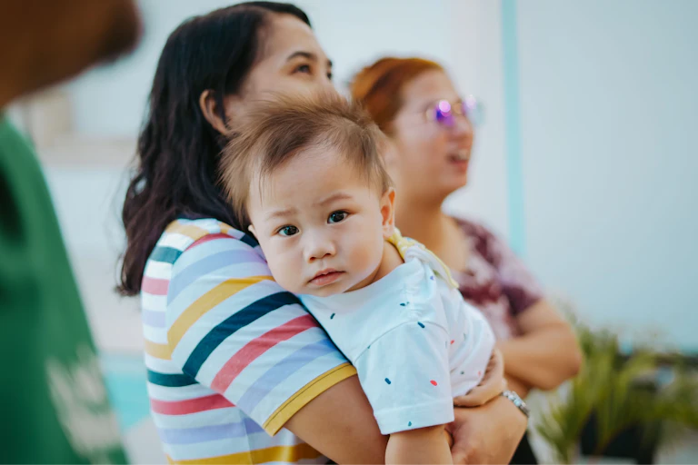 A woman holds a baby in her arms. Another woman stands in the background.