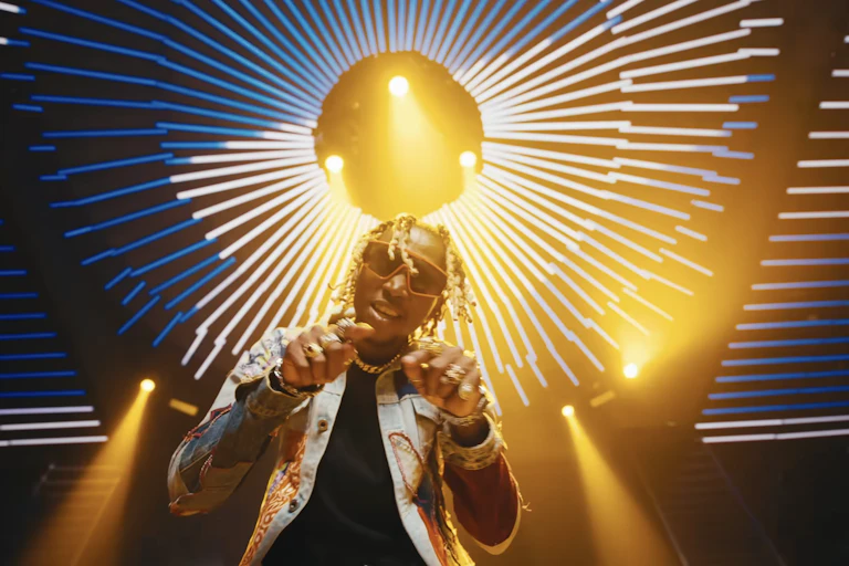 A young African man fashionably dressed with shades and peroxided dreads points both hands to camera, behind him a dazzling nightclub light.