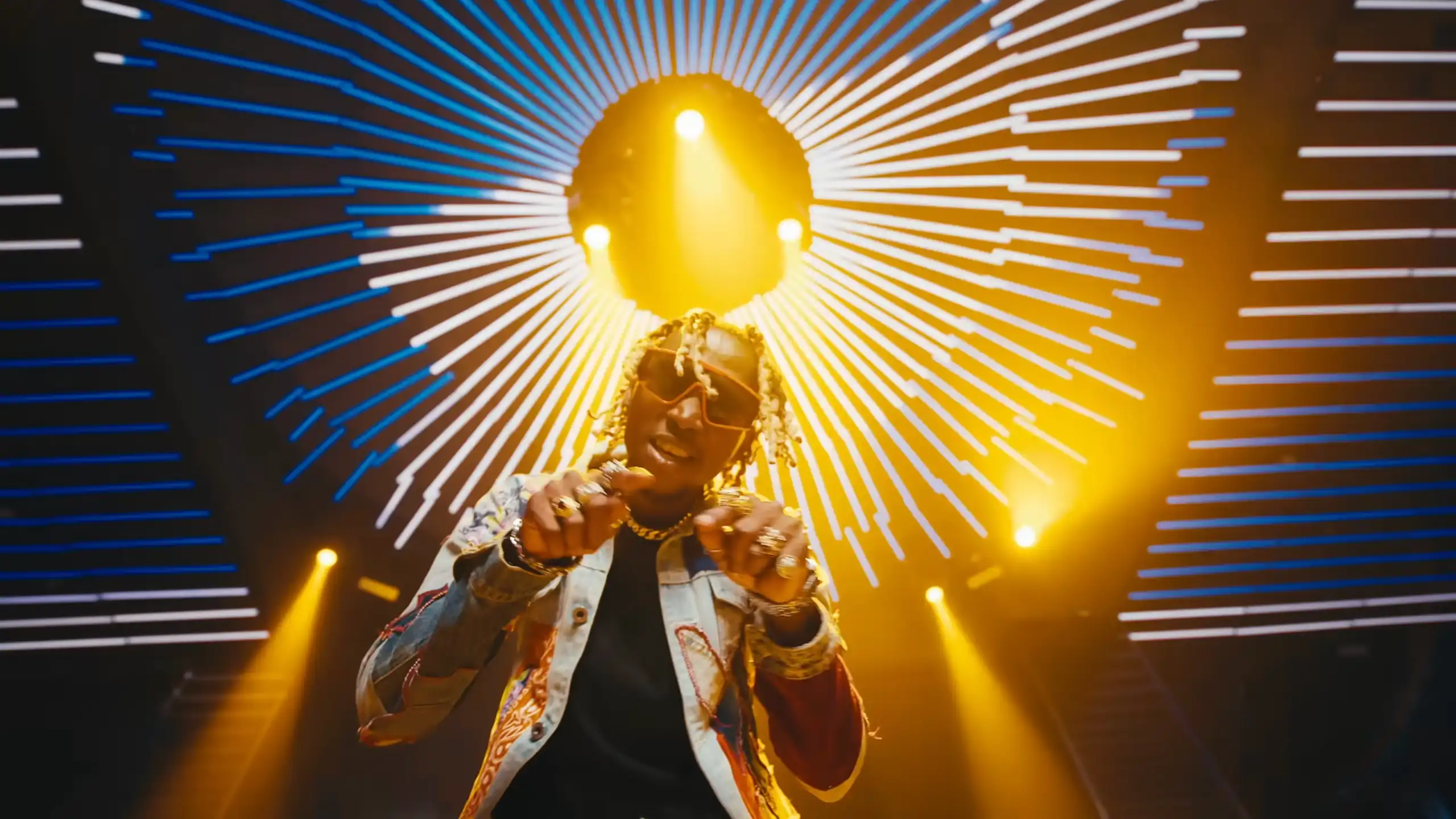 A young African man fashionably dressed with shades and peroxided dreads points both hands to camera, behind him a dazzling nightclub light.