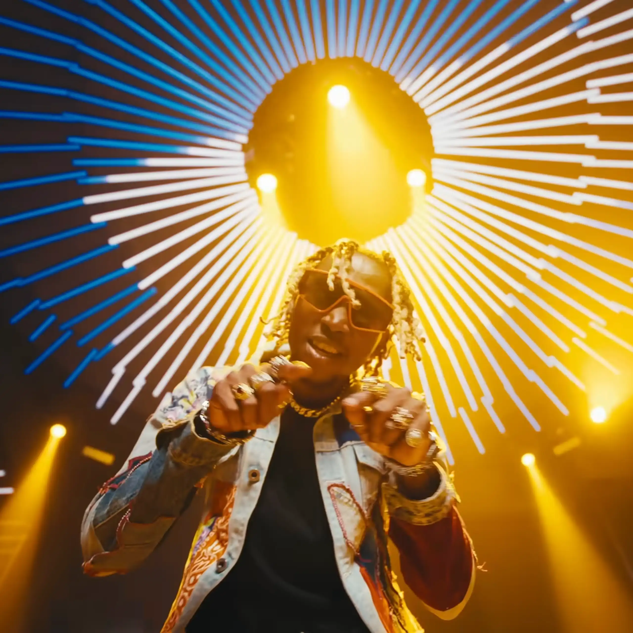 A young African man fashionably dressed with shades and peroxided dreads points both hands to camera, behind him a dazzling nightclub light.