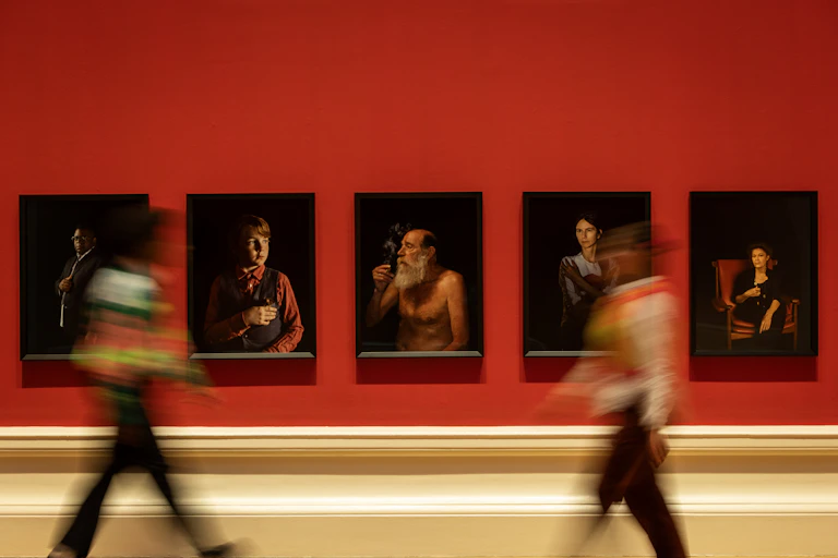 An art gallery with red walls, portraits and two blurred people walking past.