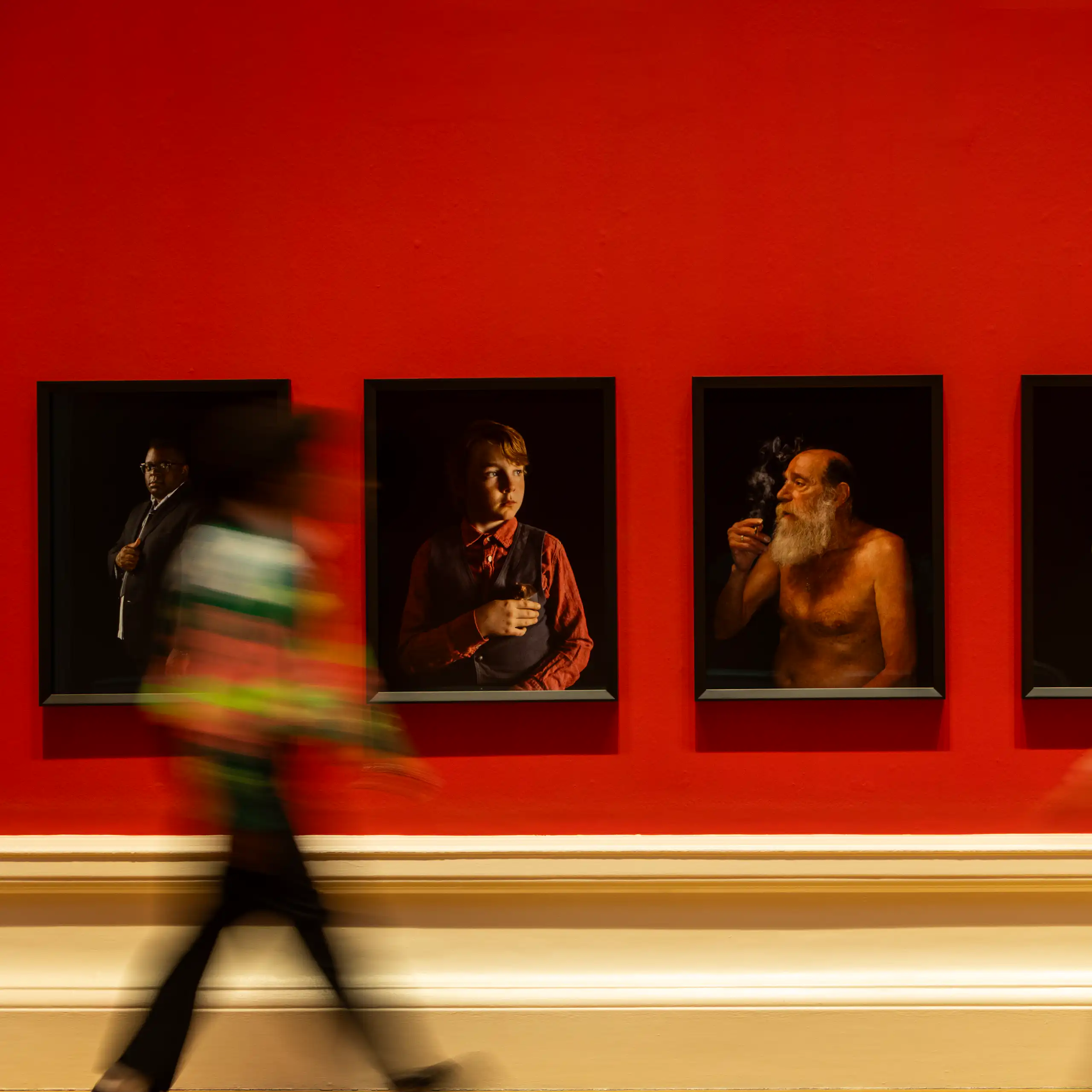 An art gallery with red walls, portraits and two blurred people walking past.