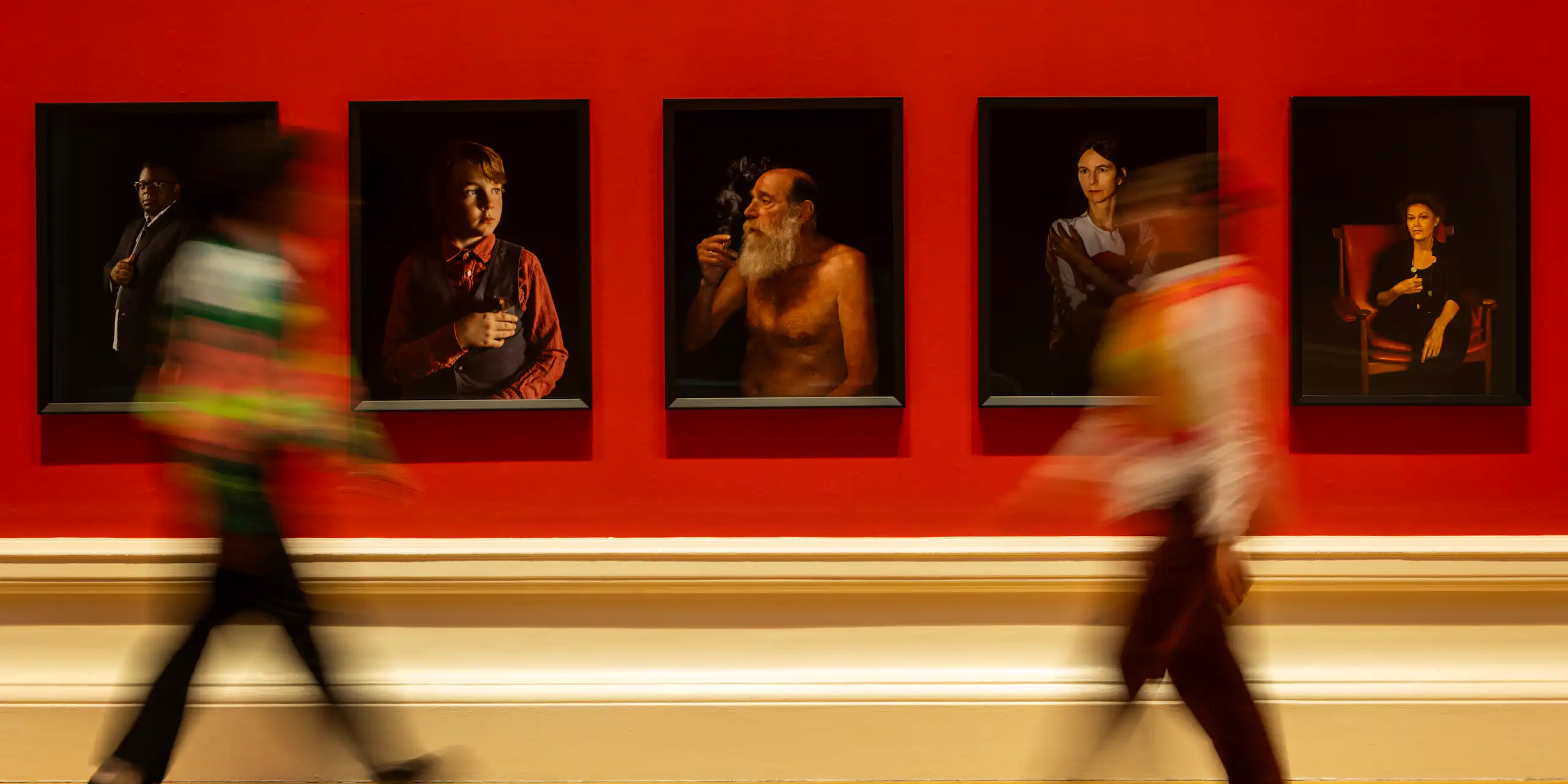 An art gallery with red walls, portraits and two blurred people walking past.