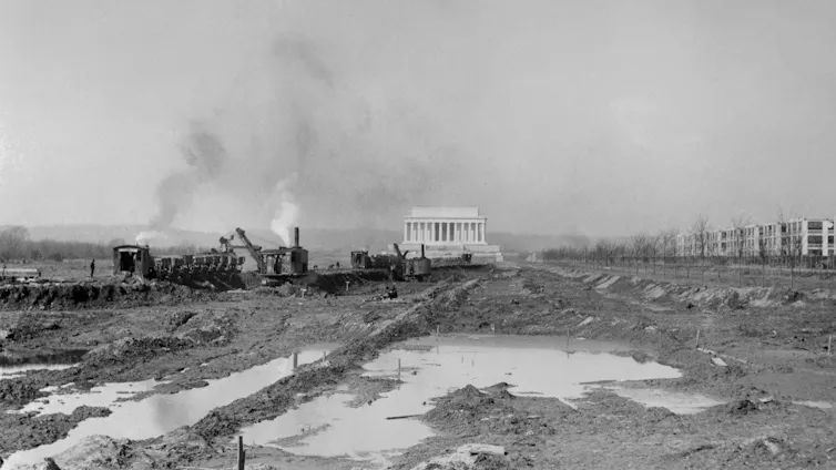 Black and white photo of ripped up land and an excavation crew in front of a large white monument.