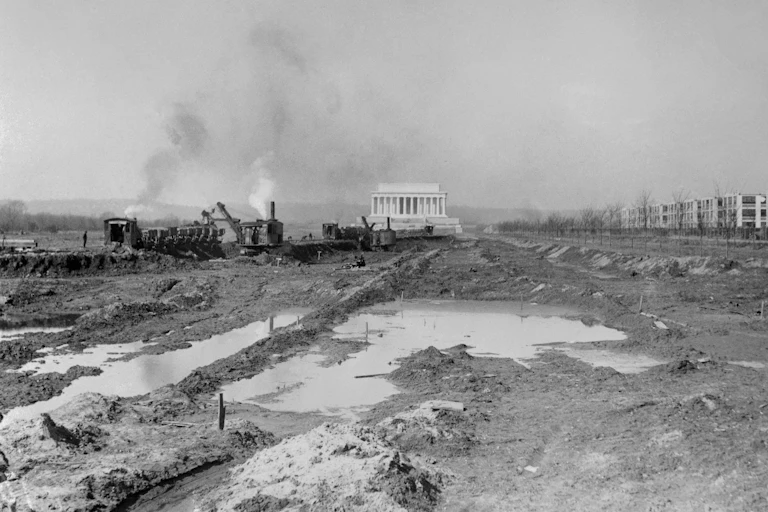Black and white photo of ripped up land and an excavation crew in front of a large white monument.