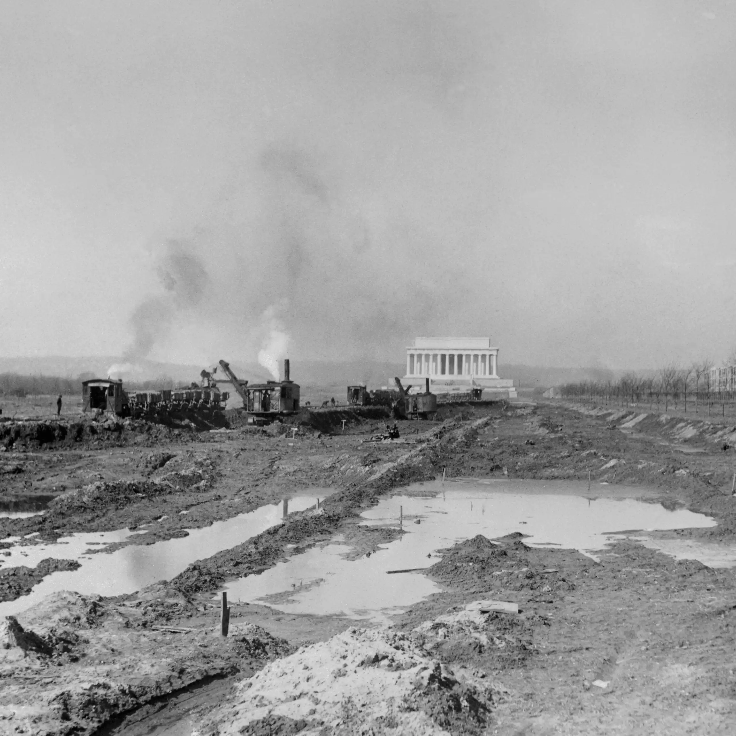 Black and white photo of ripped up land and an excavation crew in front of a large white monument.