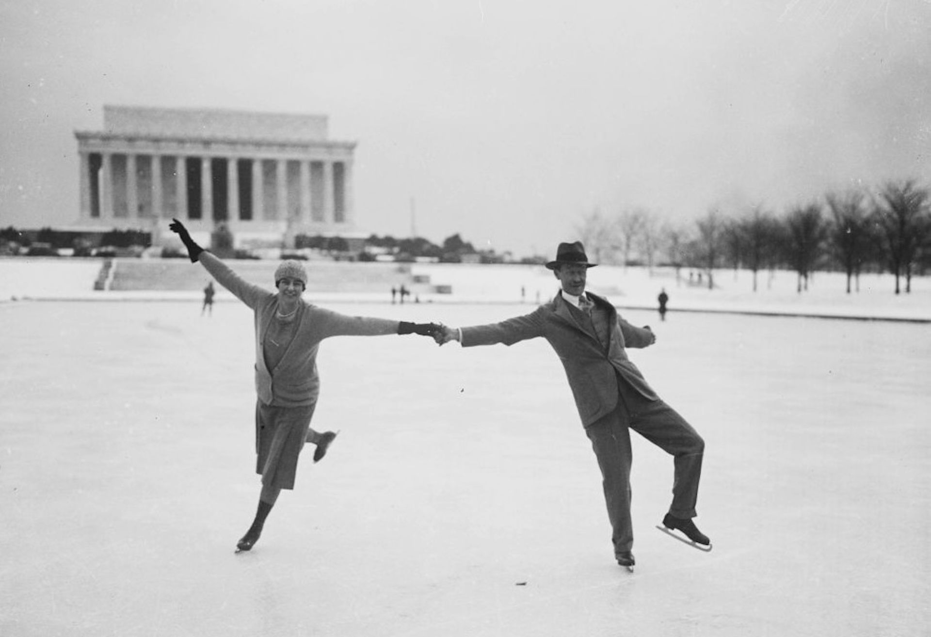 Black and white photo of a young couple holding hands, ice skating and jubilantly stretching out their arms.
