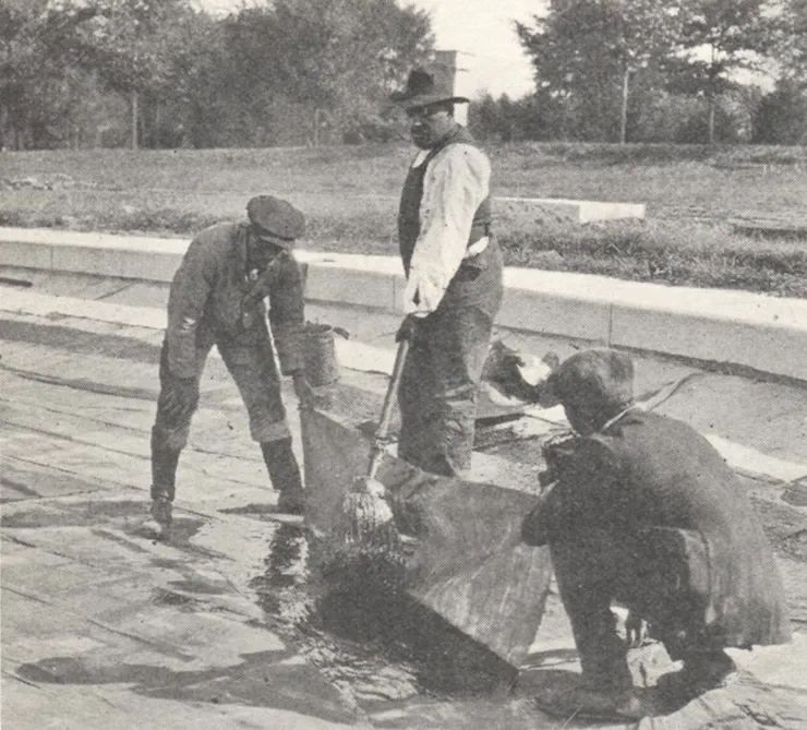 Black and white photo of three workers – two crouching, one standing – laying material on the floor of a pool under construction.