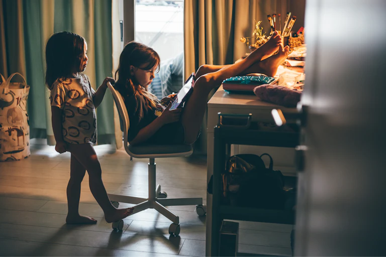 One young girl sits in a chair and puts her feet up on a desk in a dimly lit room as she looks at a screen illuminated in her hands. Another young girl stands behind her and also looks at the screen.