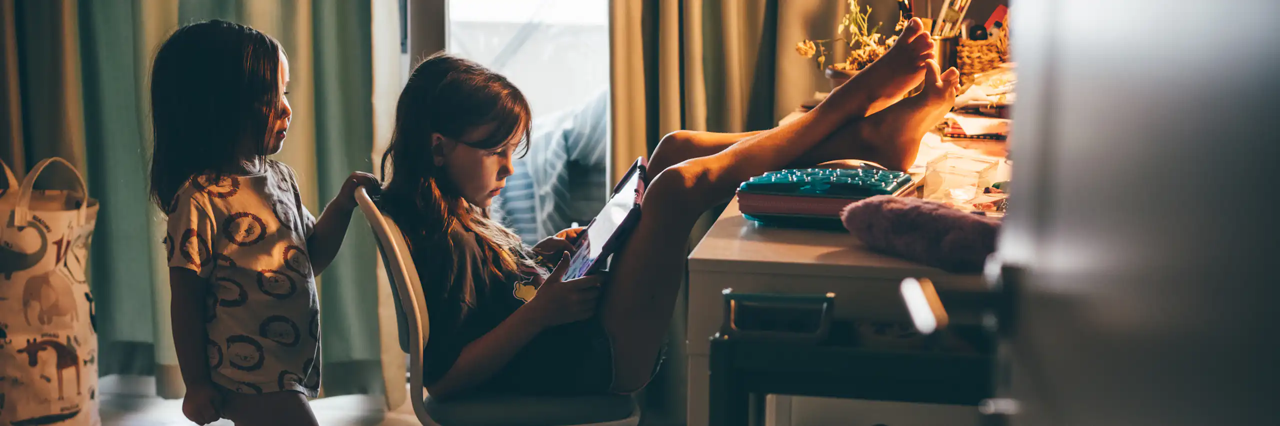 One young girl sits in a chair and puts her feet up on a desk in a dimly lit room as she looks at a screen illuminated in her hands. Another young girl stands behind her and also looks at the screen.