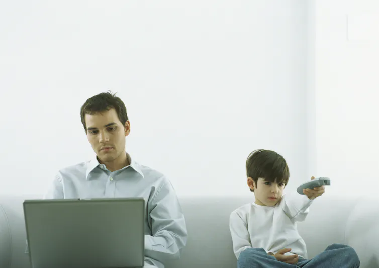 A man sits in front of a computer screen while a child holding a remote control sits next to him.