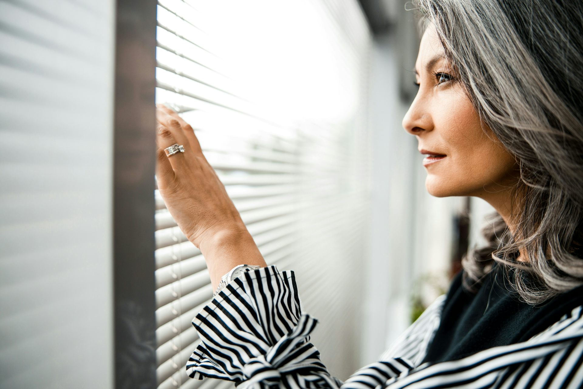 A woman with shoulder-length grey hair smiles faintly as she looks through the blinds at a window.