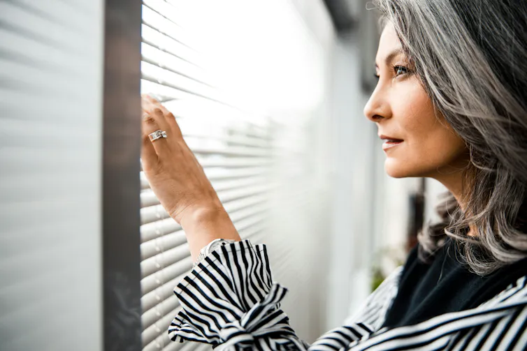 A woman with shoulder-length grey hair smiles faintly as she looks through the blinds at a window.