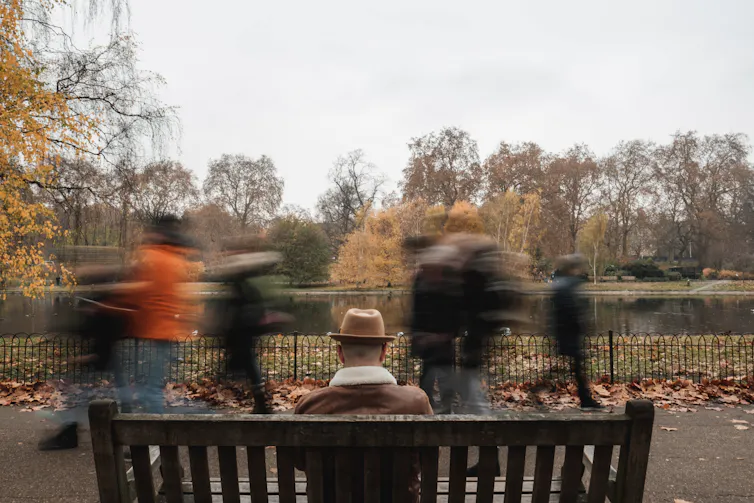 Seated man on bench in focus from behind with blurred people walking past