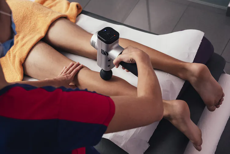 Professional physiotherapist performs shock massage with sports pistol in gym medical office.