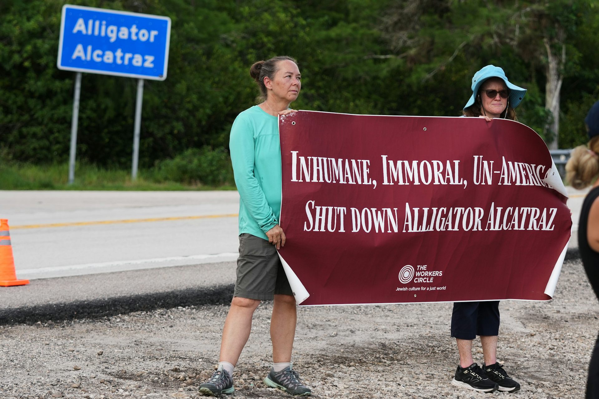 Two women hold a poster that says 'Inhumane, Immoral, Un-American, Shut Down Alligator Alcatraz'