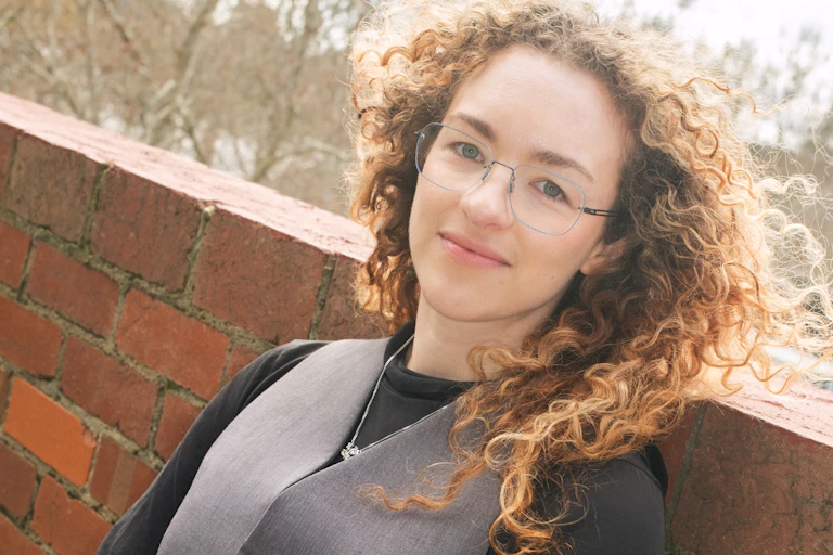 The author of Wormhole: a young white woman with curly hair and glasses, wearing a grey vest over a long-sleeved black top.