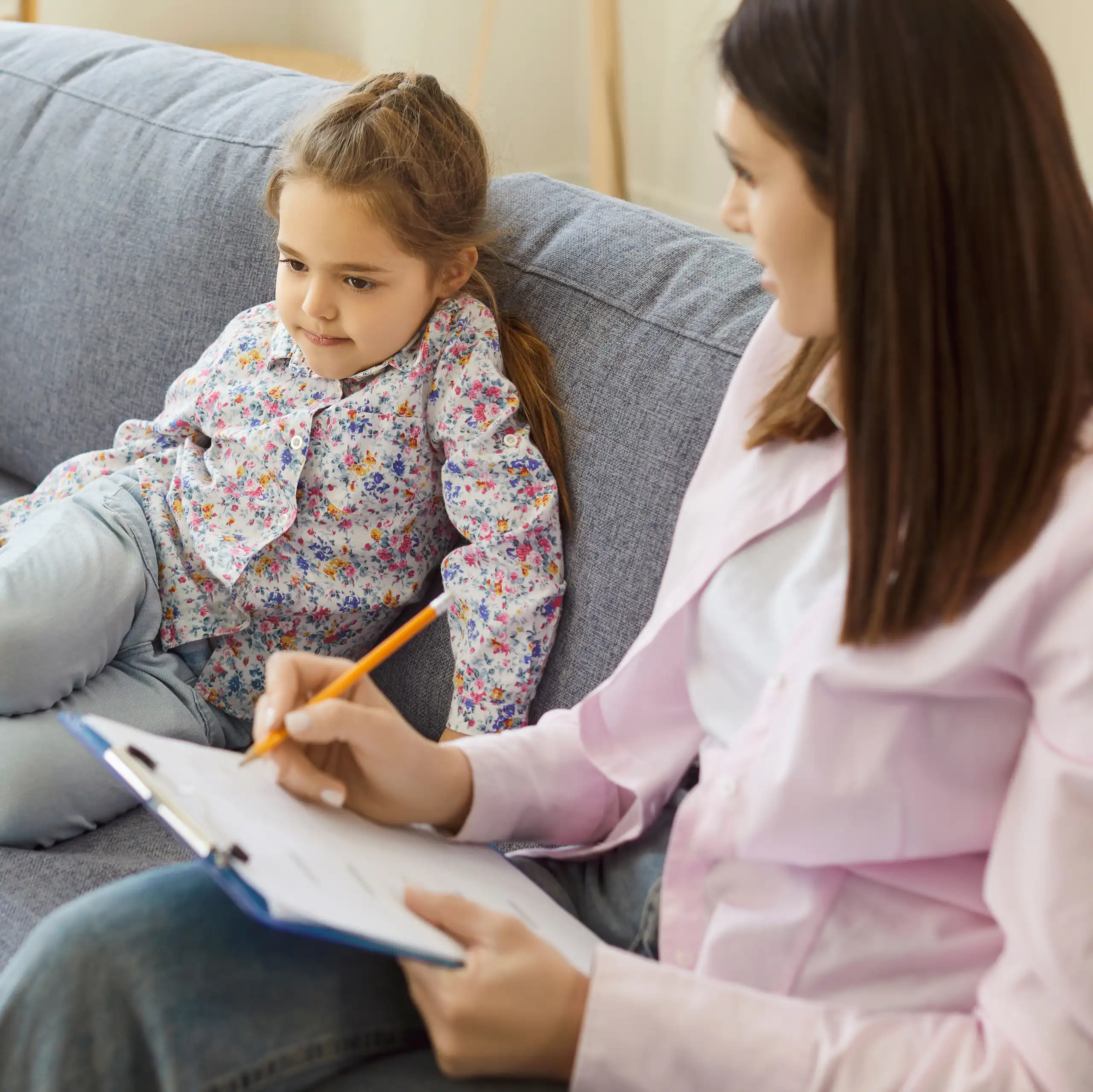 A young girl sits on a couch next to a female psychiatrist, who is taking notes on a clipboard.