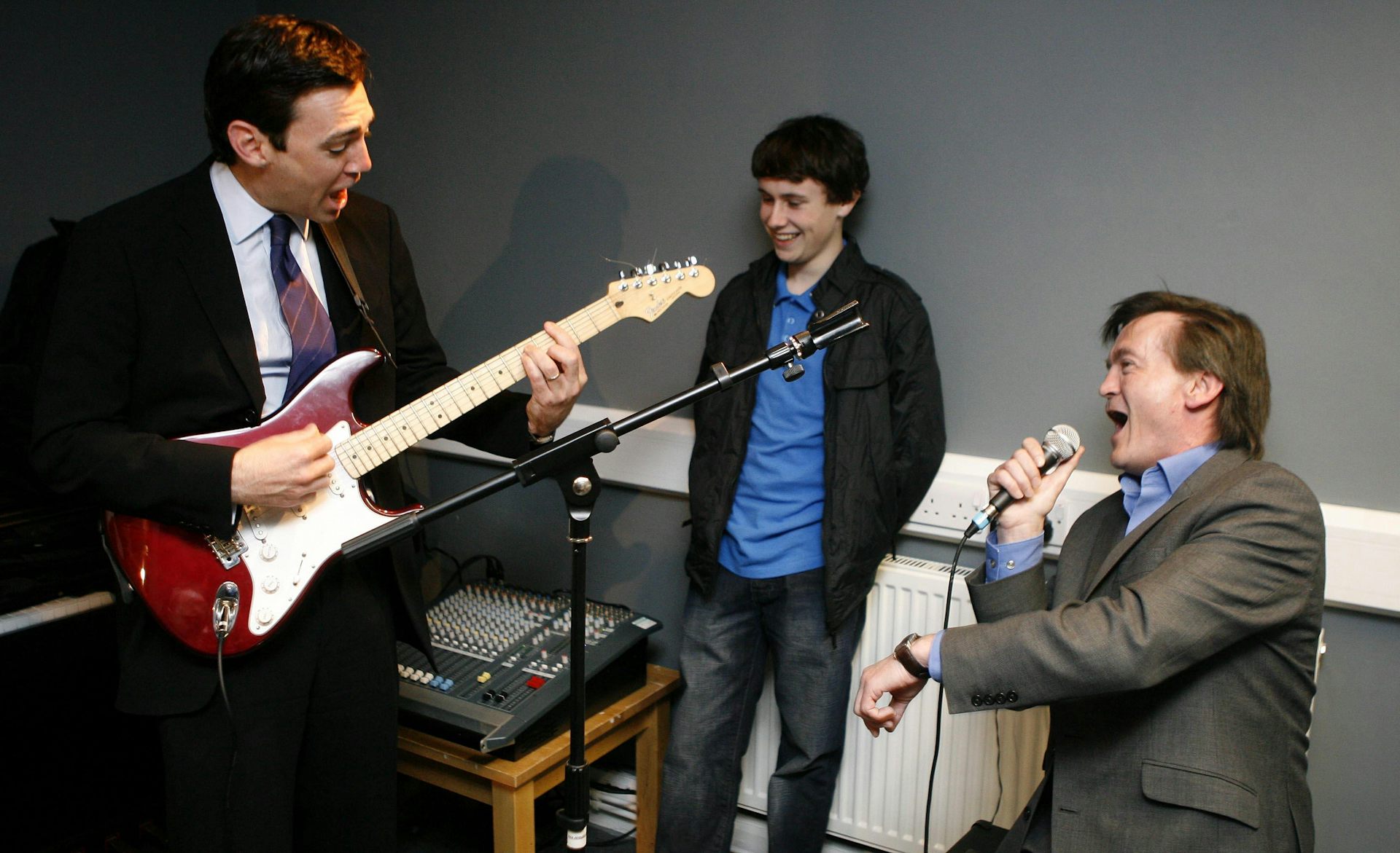 Two older men one with a guitar, one singing and younger man looking on.