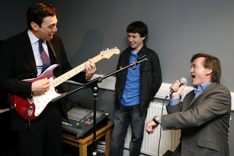 Two older men one with a guitar, one singing and younger man looking on.