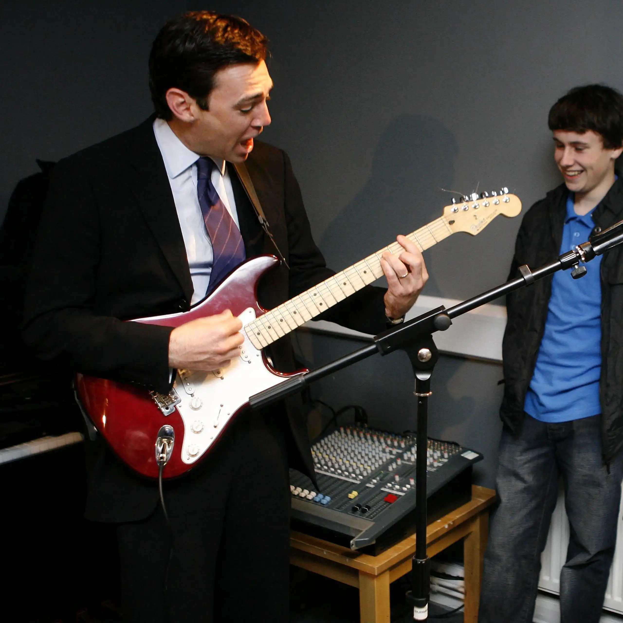 Two older men one with a guitar, one singing and younger man looking on.