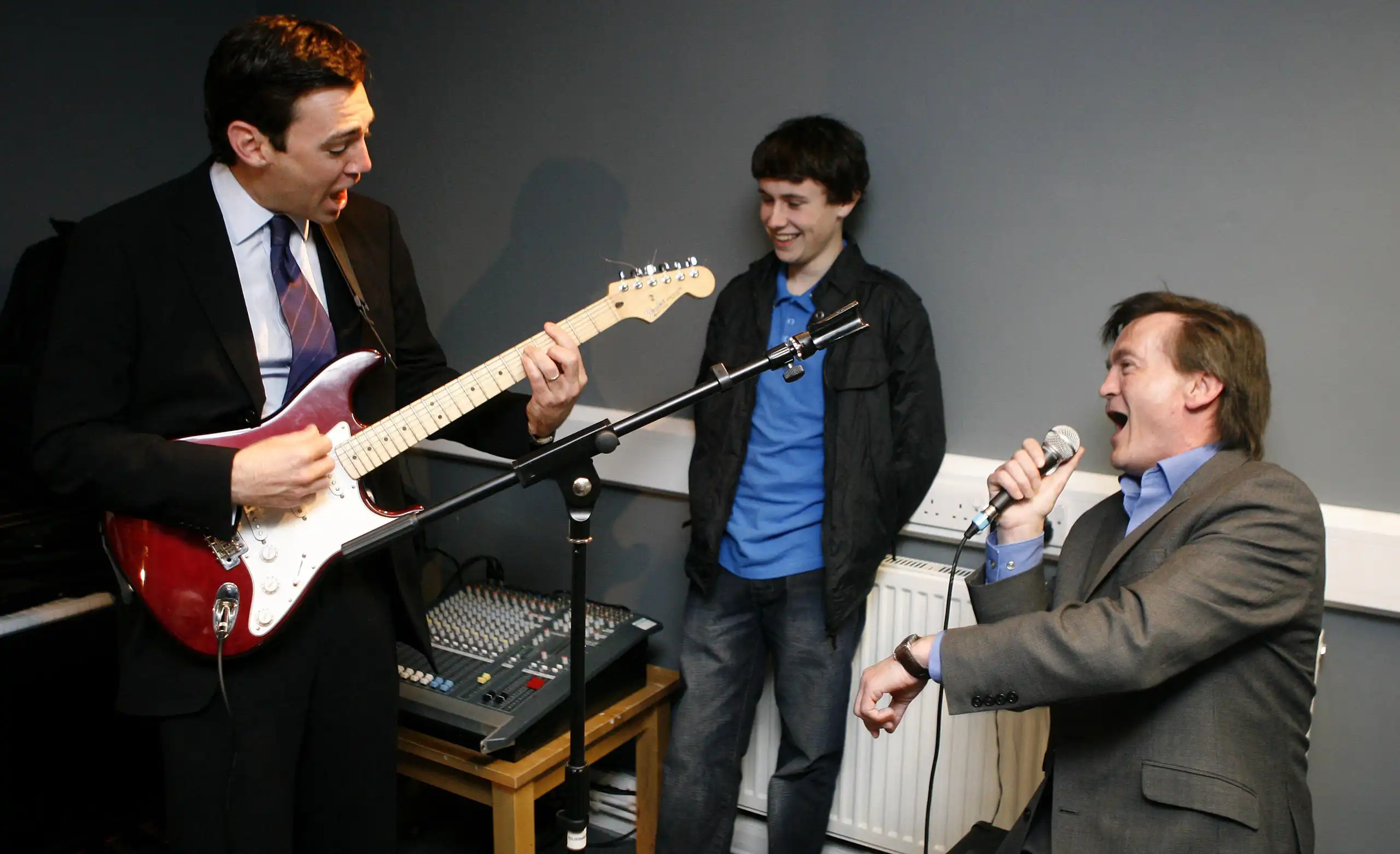 Two older men one with a guitar, one singing and younger man looking on.
