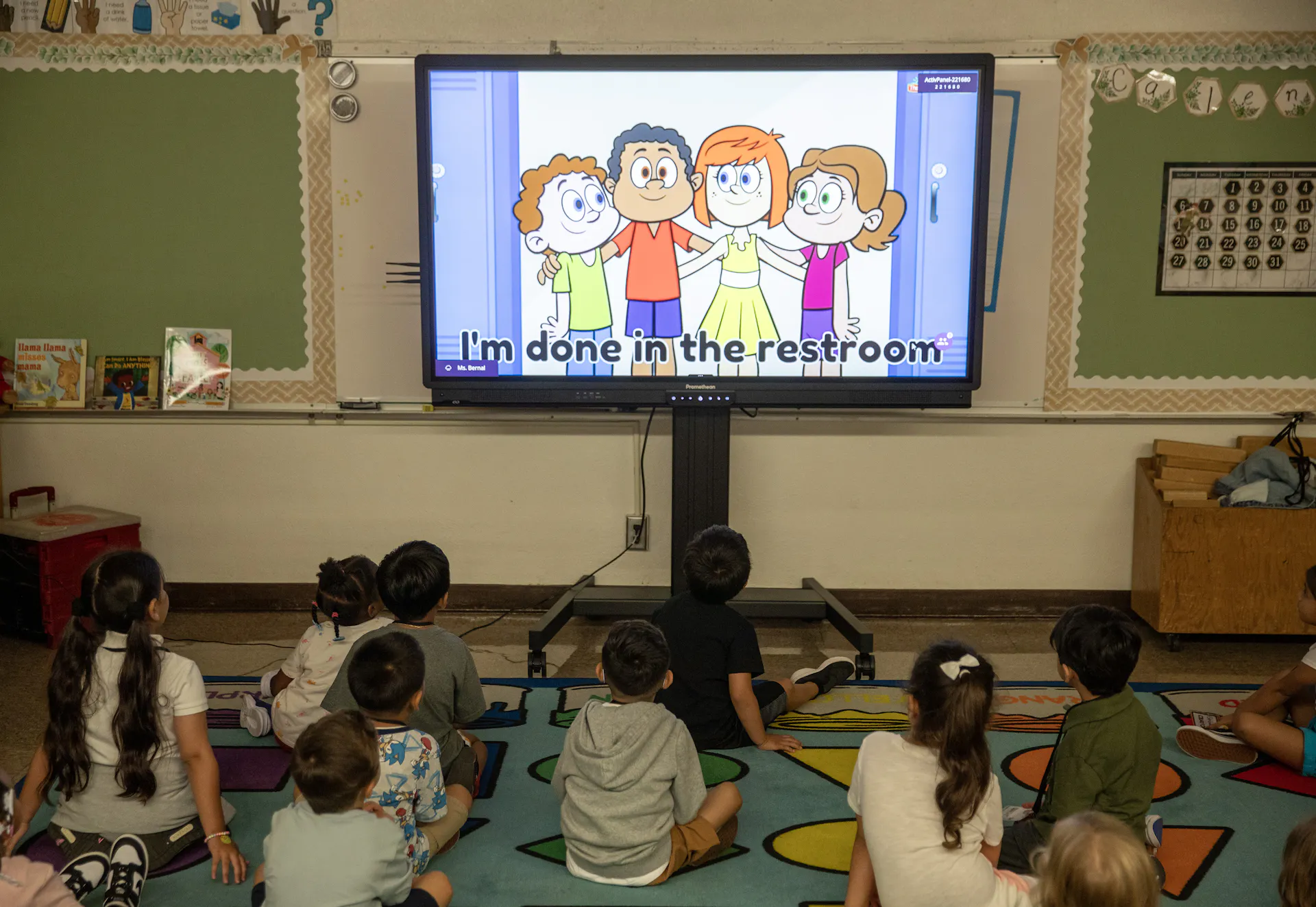 A group of children sit on the floor looking at a screen that shows cartoon characters and the words 'I'm done in the restroom.'
