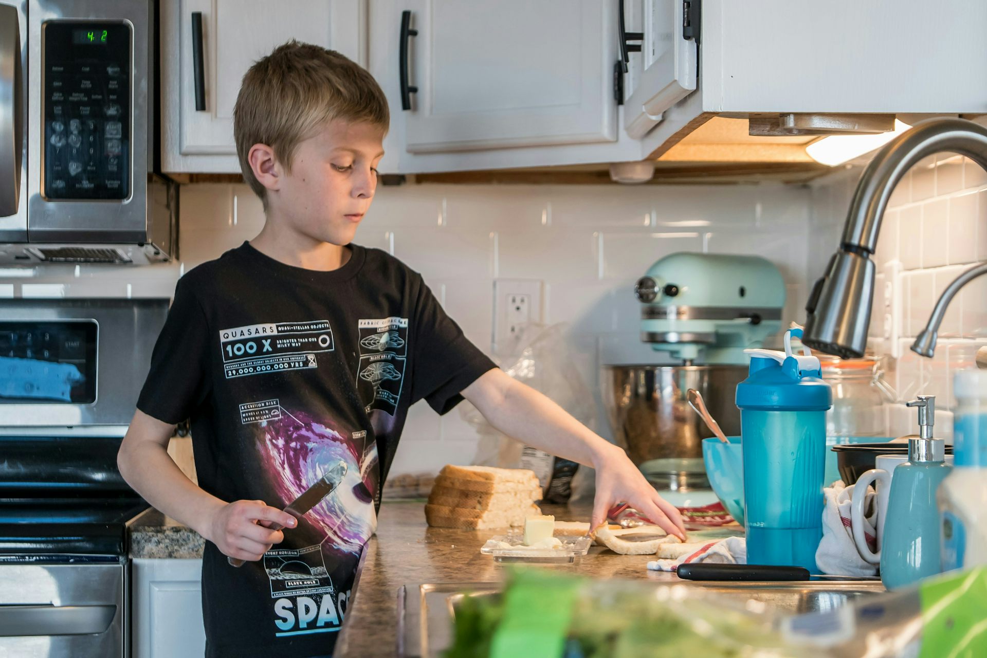 A boy in a t-shirt buttering bread at a kitchen counter