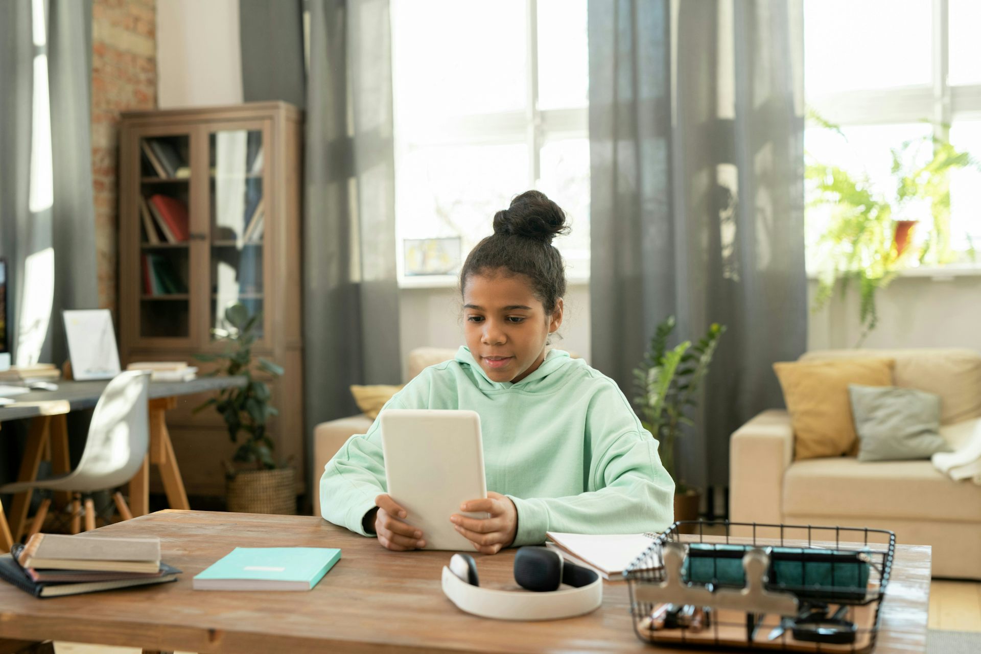 A girl in a green hoodie alone in a living room, sitting at a table looking at a tablet