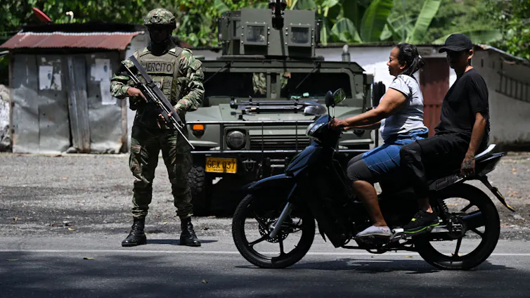 Two people on a motorbike ride past a man in army uniform carrying a large gun.